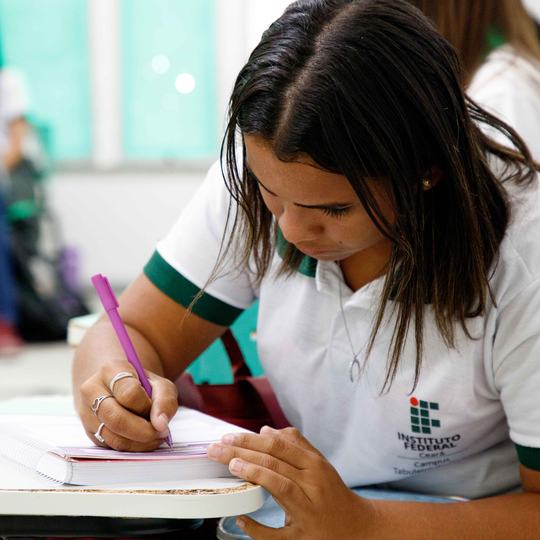 Estudante com farda do IFCE, debruçada sobre o caderno fazendo uma anotação.