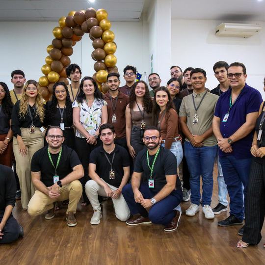 Estudantes e professores do campus de Quixadá estão olhando para a câmera em cima do palco do auditório. Ao fundo, balões dourado formam um arco.