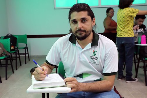 Estudante do curso de Letras, usando a farda do IFCE, sentando na carteira, segurando uma caneta e anotando no caderno.