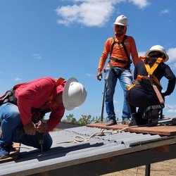 Três estudantes utilizando equipamentos de segurança, sendo dois deles agachados, realizando a instalação de placas solares, sendo que um deles está com uma furadeira em mãos.
